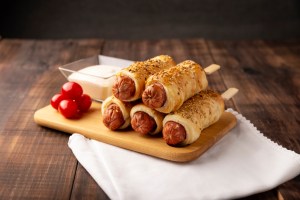 close up of delicious snack of sausages wrapped in puff pastry with sesame seeds accompanied by cherry tomatoes and sauce. Served on wooden board